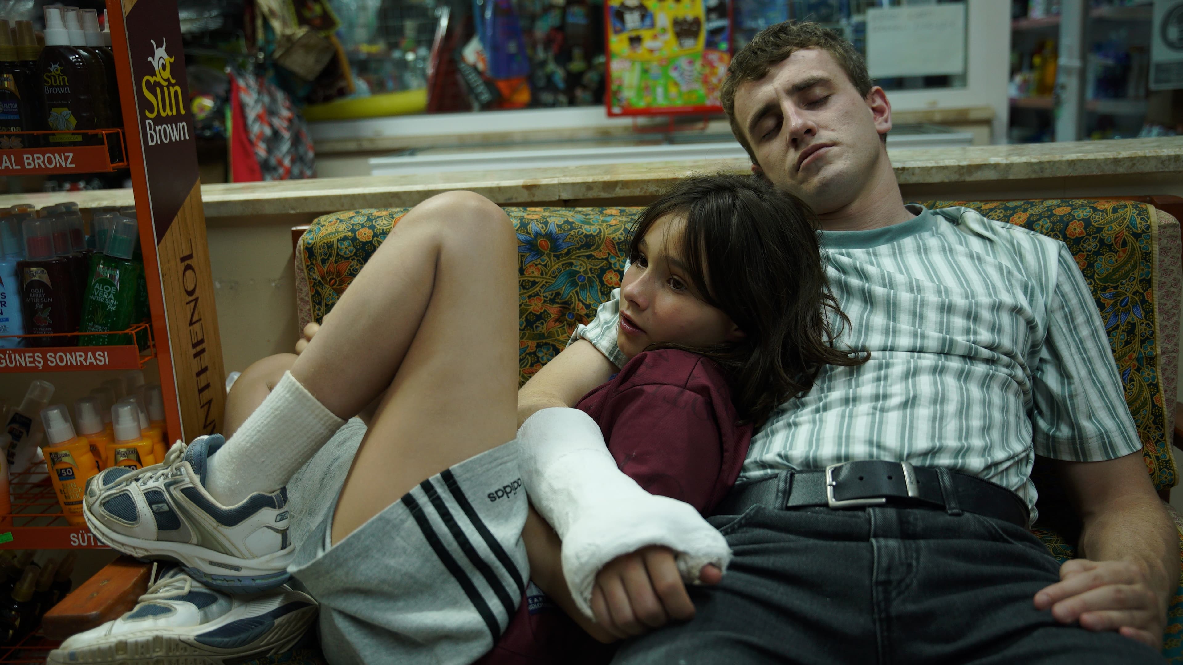A young girl is held in the arms of a young man with a cast on his lower right arm in this still from Aftersun.
