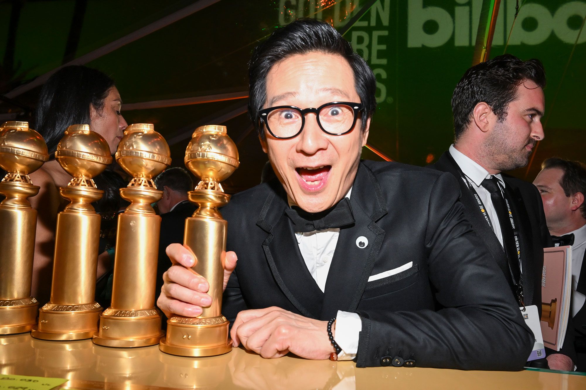 Actor Ke Huy Quan, wearing a black and white tuxedo with black rimmed glasses, smiles while gripping a Golden Globe award.