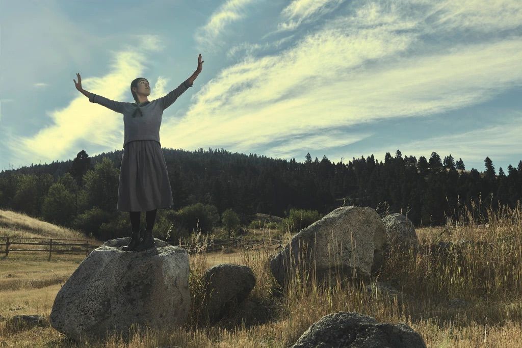 Aminah Nieves' 1923 character Teonna stands atop a large rock in a field in this still from the show.